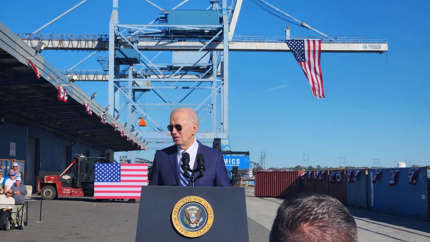 Public official speaking at a port with a container crane and American flag behind