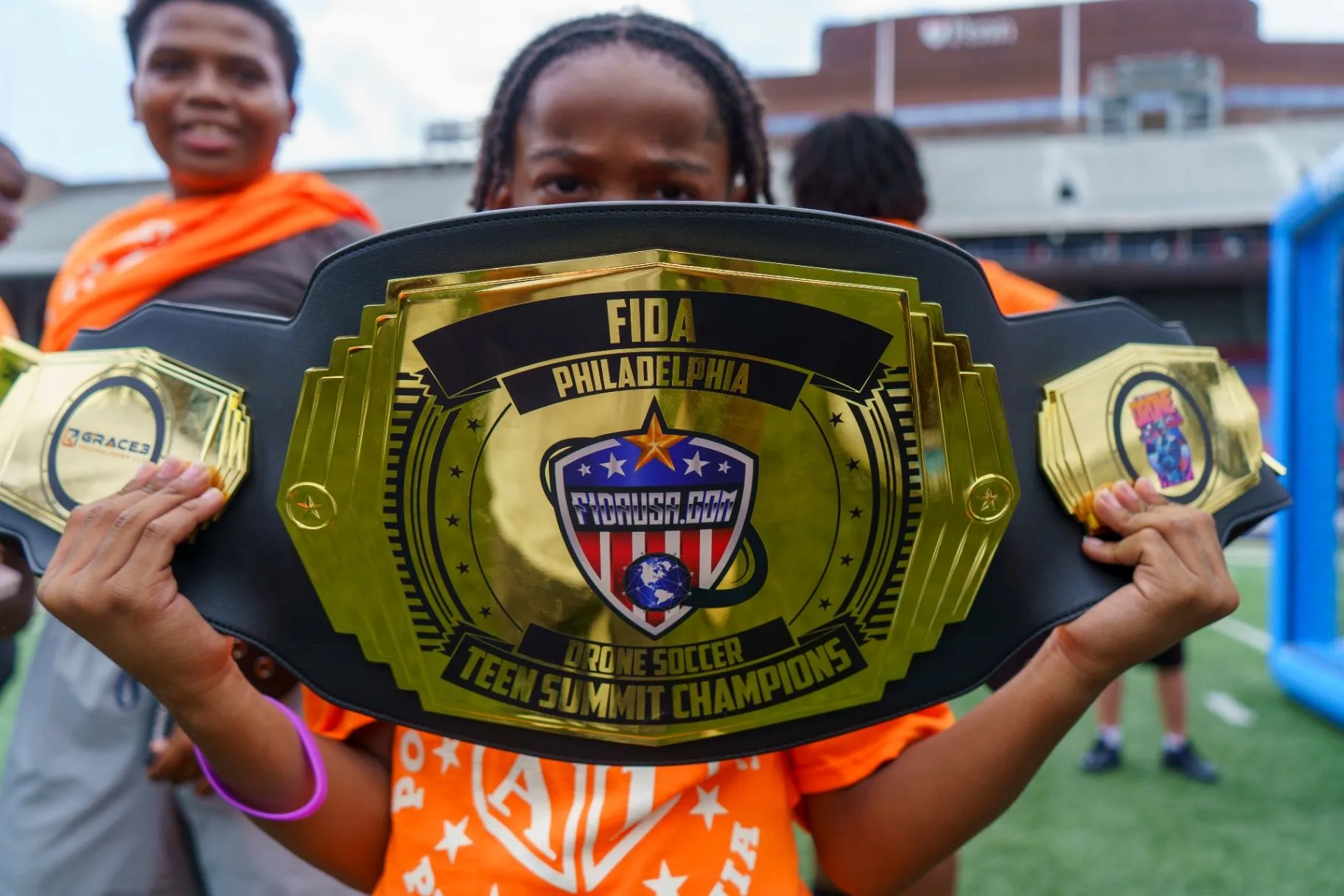 A student holding up a large gold championship belt reading FIDA Philadelphia Drone Soccer Teen Summit Champions
