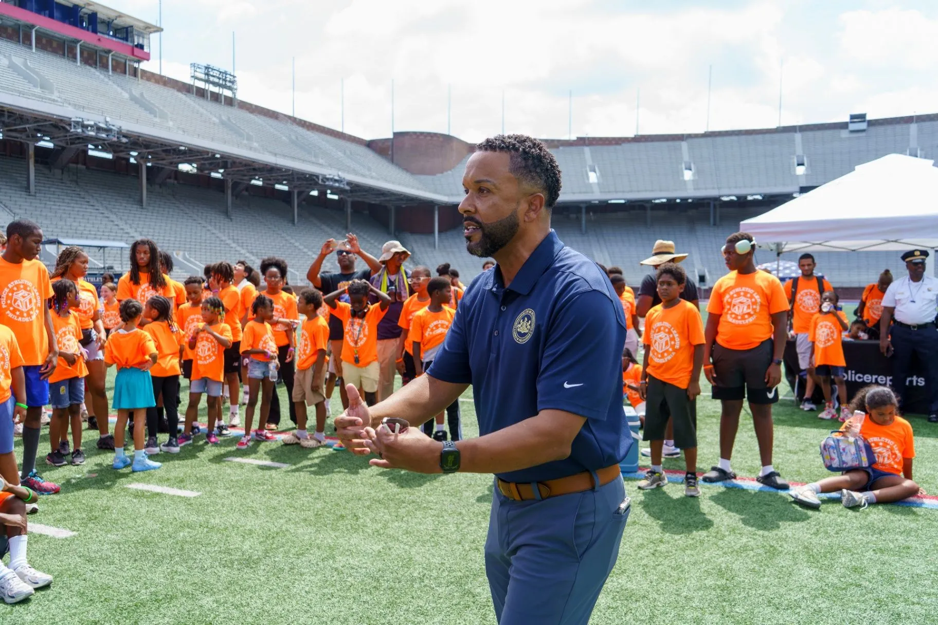 A speaker in a navy polo addressing students in orange shirts on the field inside a stadium