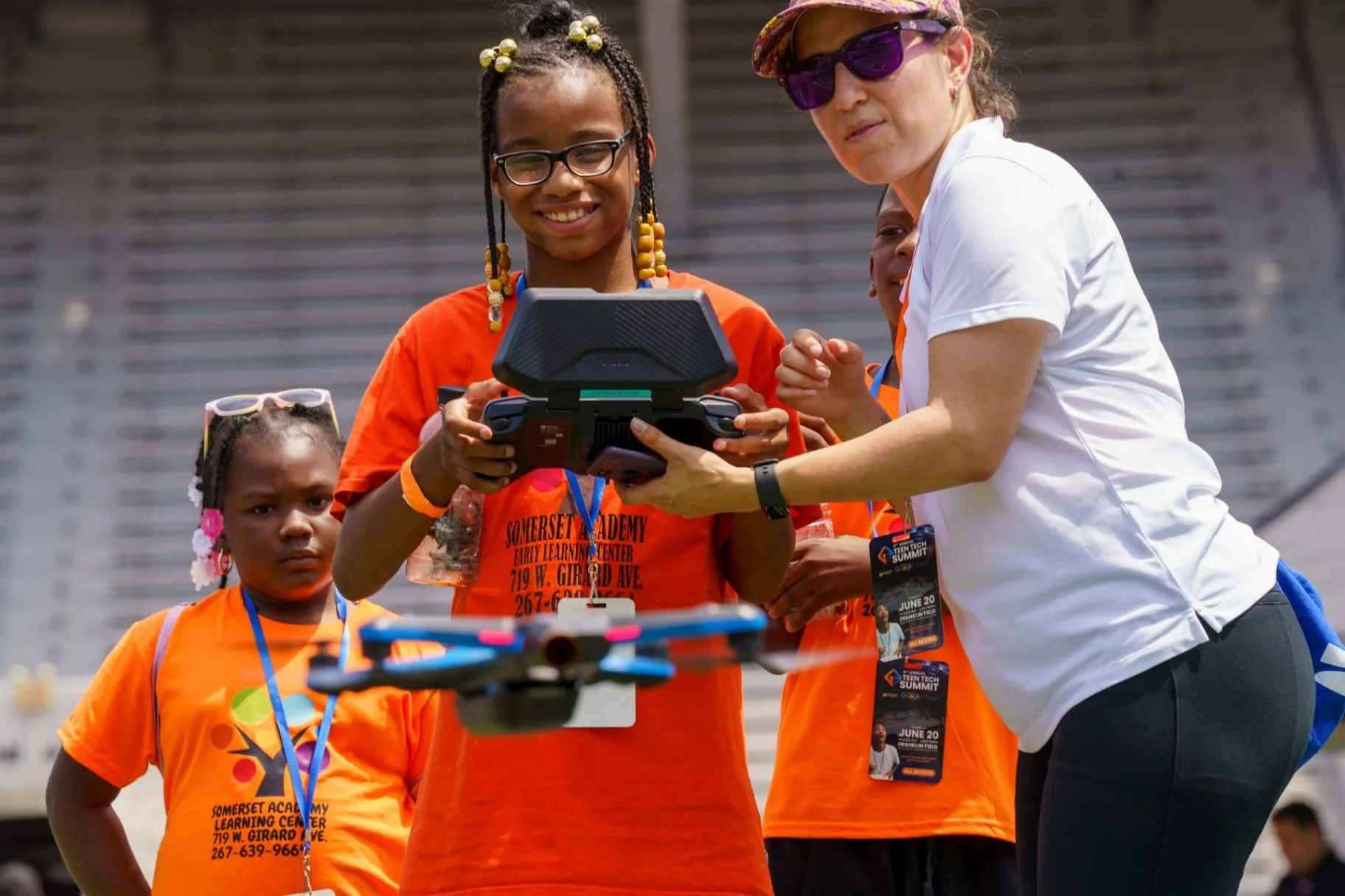 A student in an orange Somerset Academy shirt learning to fly a drone while an instructor guides her hands at Franklin Field