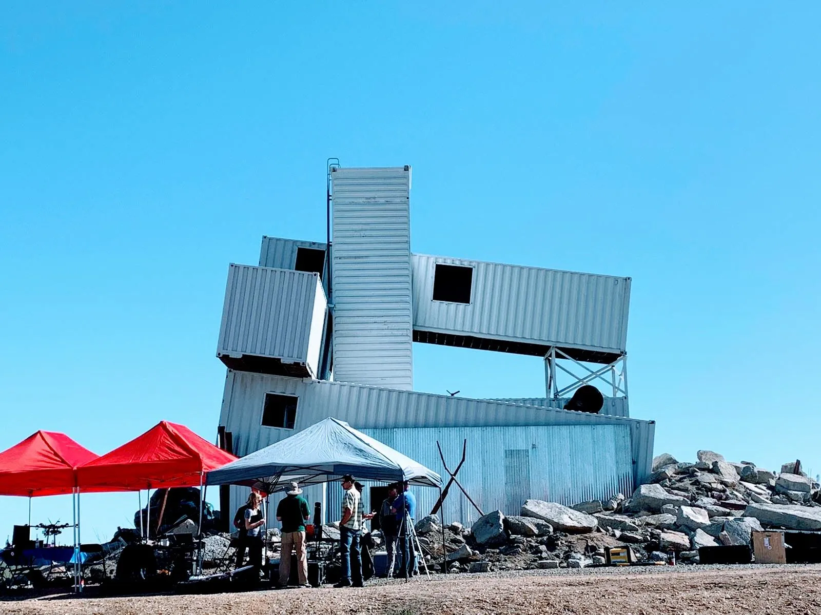 People and drone equipment gathered beneath tents beside a stacked white shipping-container training structure